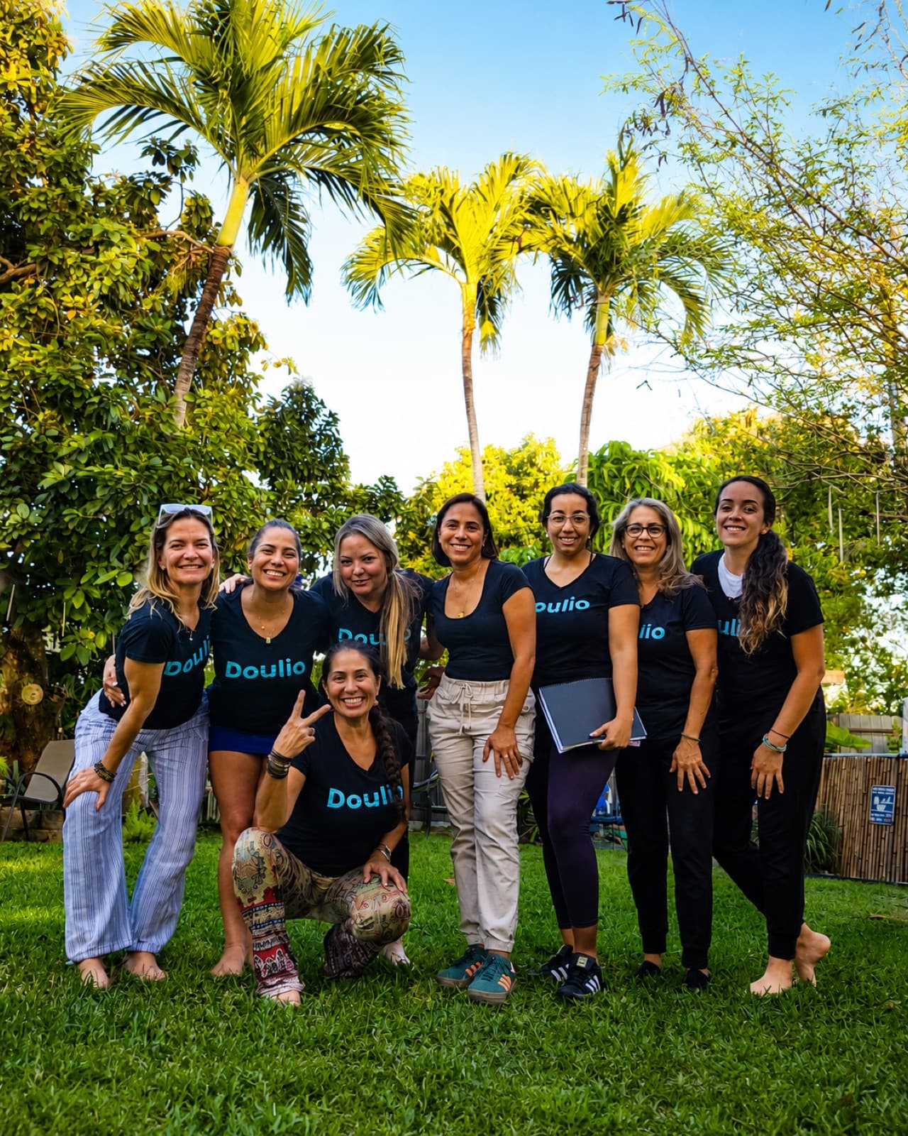 A group of Doulio trainers in matching Doulio shirts smiling together outdoors under palm trees